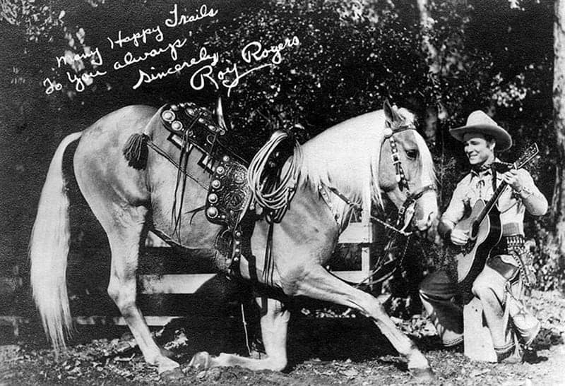 Black-and-white publicity photo of cowboy star Roy Rogers standing beside his palomino stallion Trigger